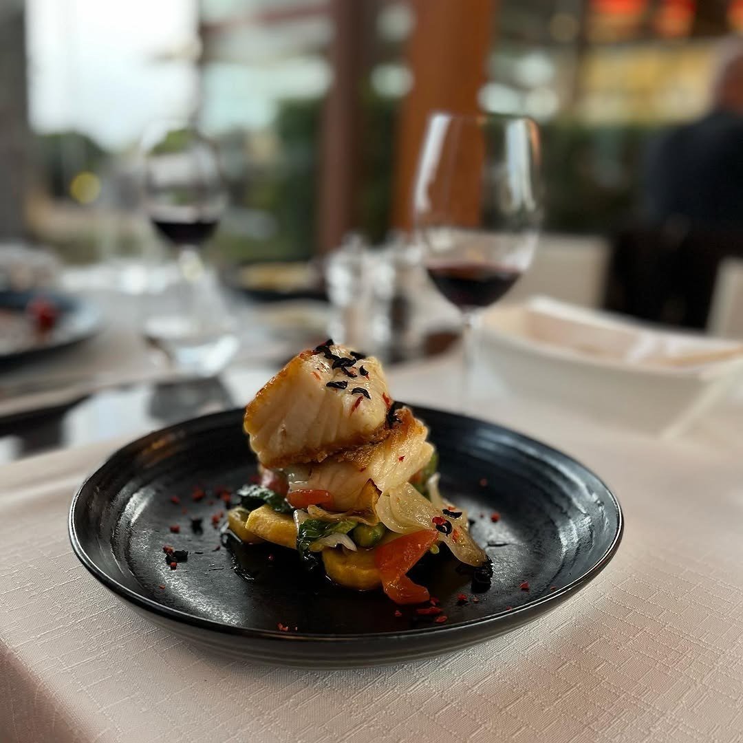 Posta de bacalhau com legumes num prato escuro, com um copo de vinho tinto atrás