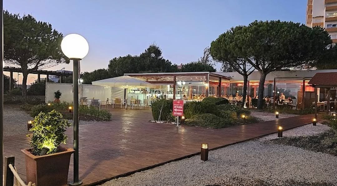 Passadiço de madeira iluminado e o restaurante envidraçado entre pinheiros, ao anoitecer